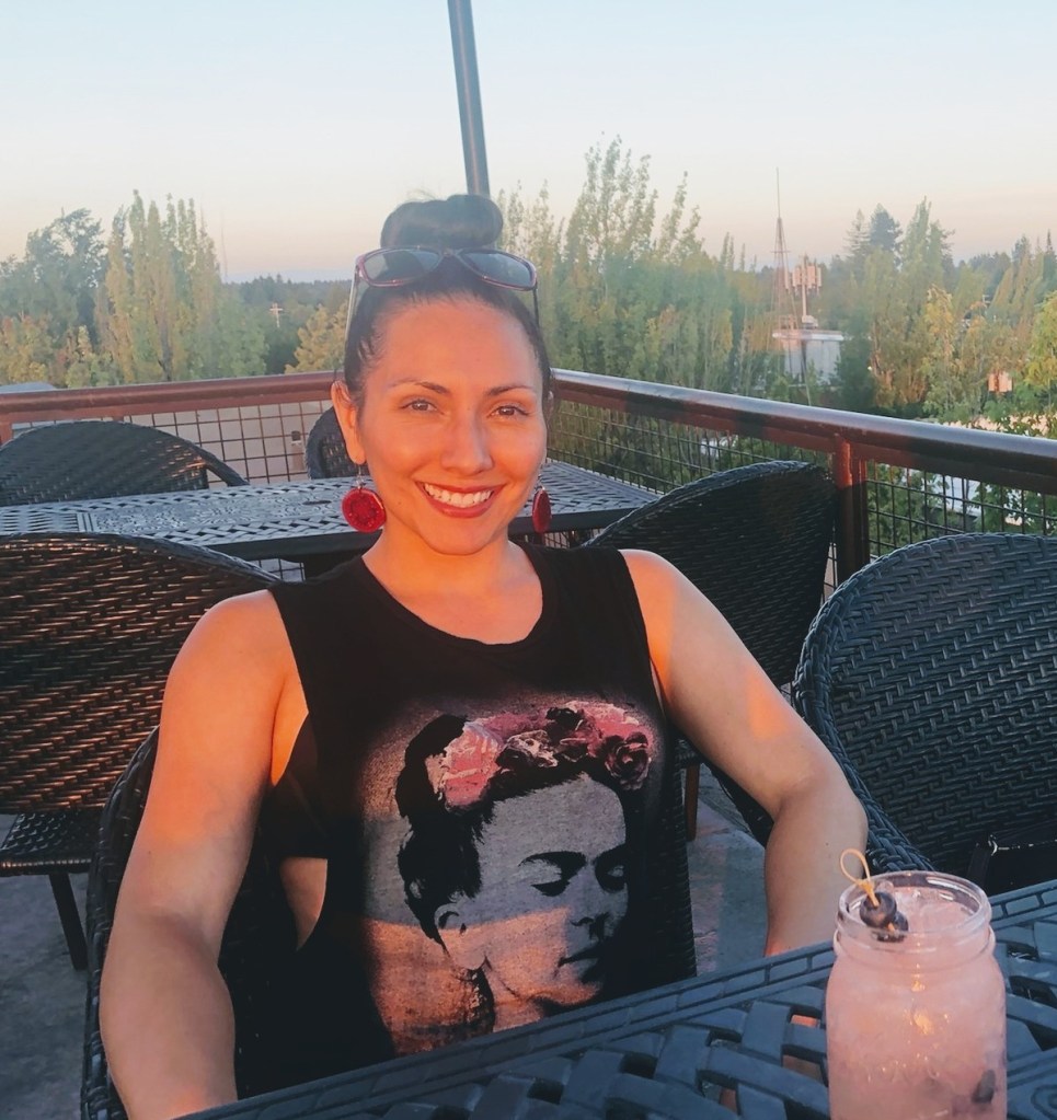 A smiling woman sits at a rooftop table, enjoying the last of the day's sunshine.