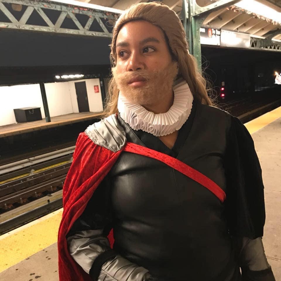 A woman is standing on a train platform dressed in black, with a red cape, white ruffle, and long blonde wig with a matching beard. She is doing cosplay as Shakespearian Thor.