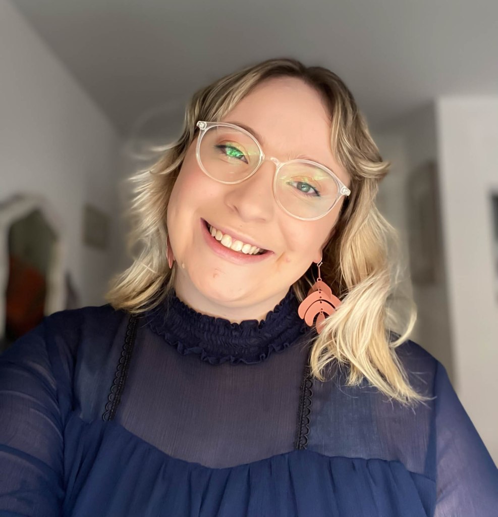 A white woman with shoulder-length blonde waves, glasses, and a navy blue shirt beams at the camera.