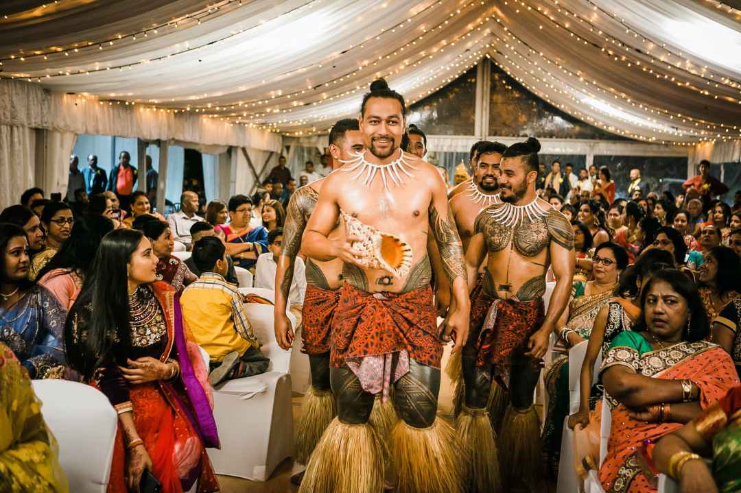 Pacific Islander men walk down an aisle preparing to dance at a wedding. The tent ceiling is lit with twinkly lights, and people in colorful Indian attire fill the rows of cloth white chairs.