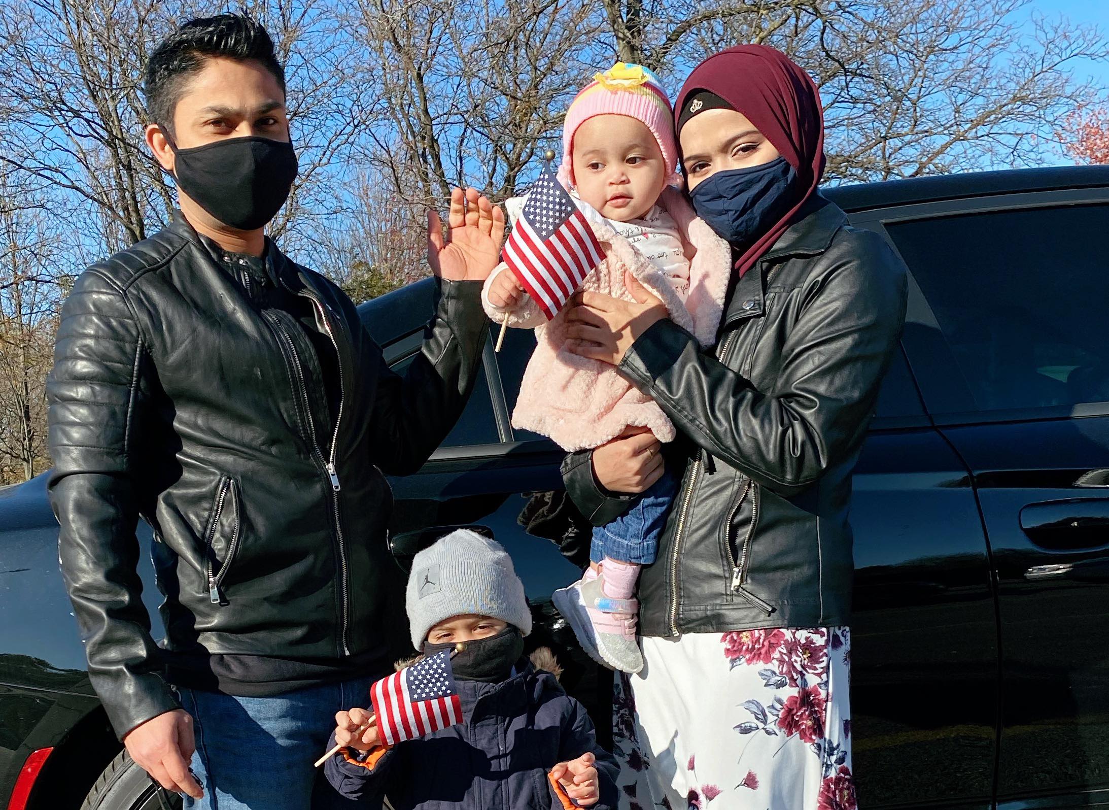 A Rohingya family in Chicago poses in front of their car. The parents wear black leather jackets and are masked. The mom is in a red hijab that matches her flowery dress. The two children wave American flags.