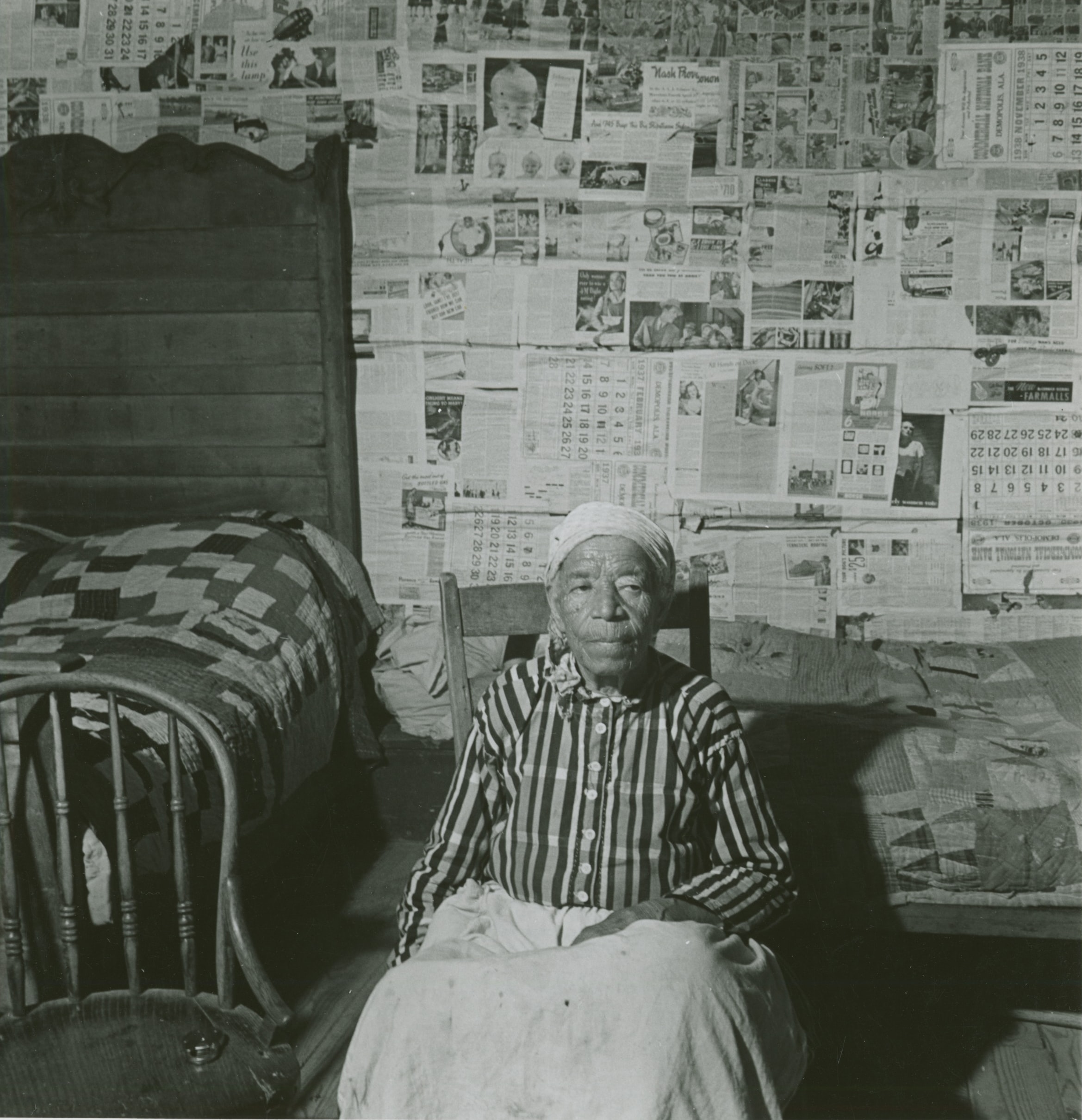 Black and white photo of an elderly mulatto woman in her house. She is sitting, and the wall behind her is lined with newspapers.