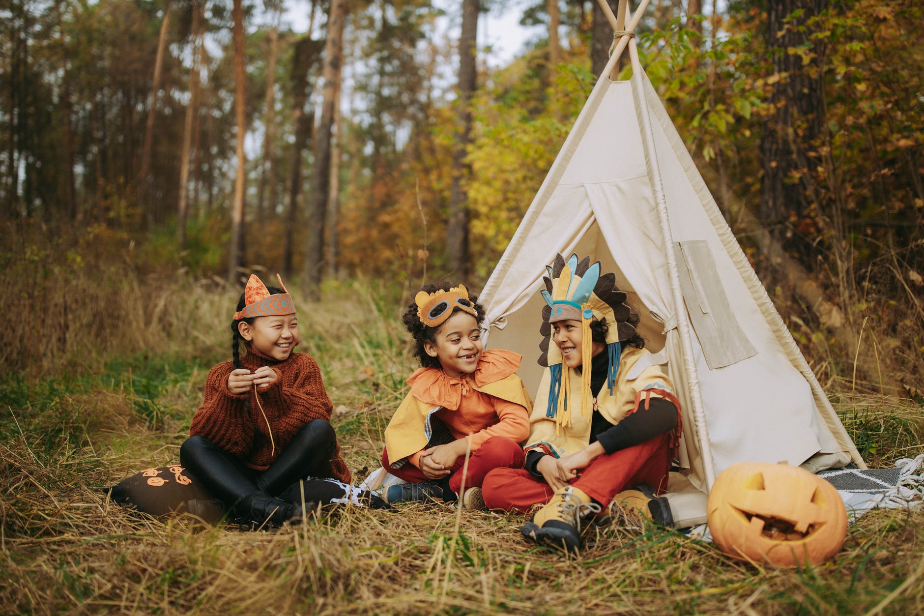 Three young children sit outside in a forested area by a teepee. It is a candid shot, and they are smiling, laughing, and wearing homemade headdresses. A carved pumpkin sits off to the right.