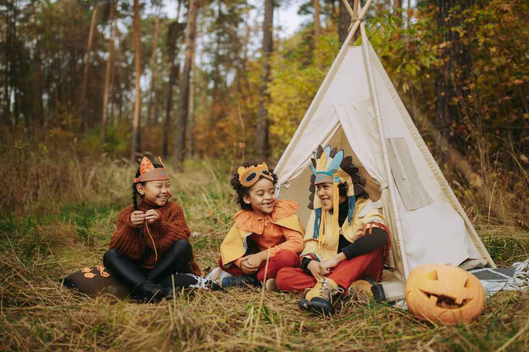 Three young children sit outside in a forested area by a teepee. It is a candid shot, and they are smiling, laughing, and wearing homemade headdresses. A carved pumpkin sits off to the right.