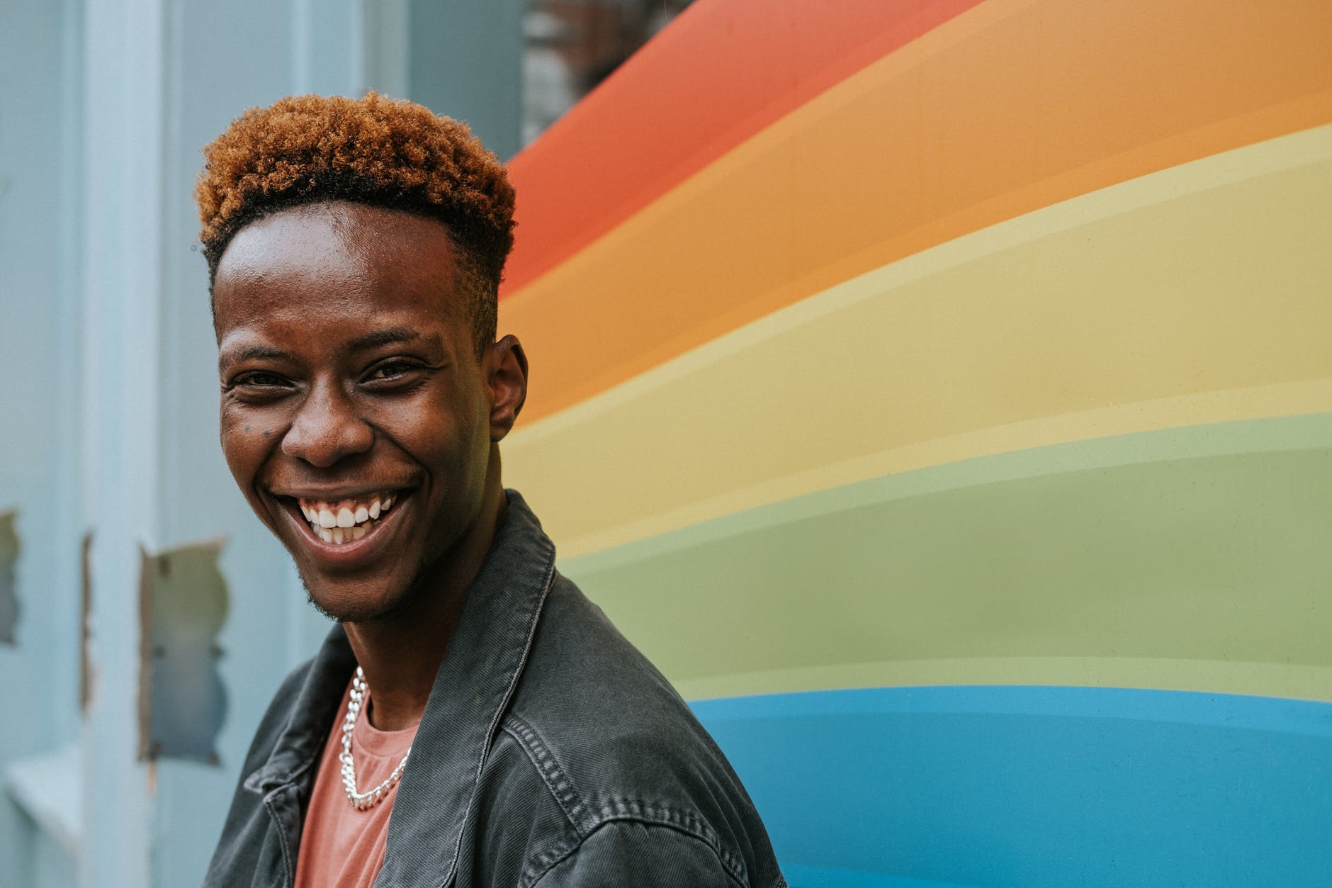 A young Black person smiles and laughs. They are offset in front of a vaguely gay flag like background.
