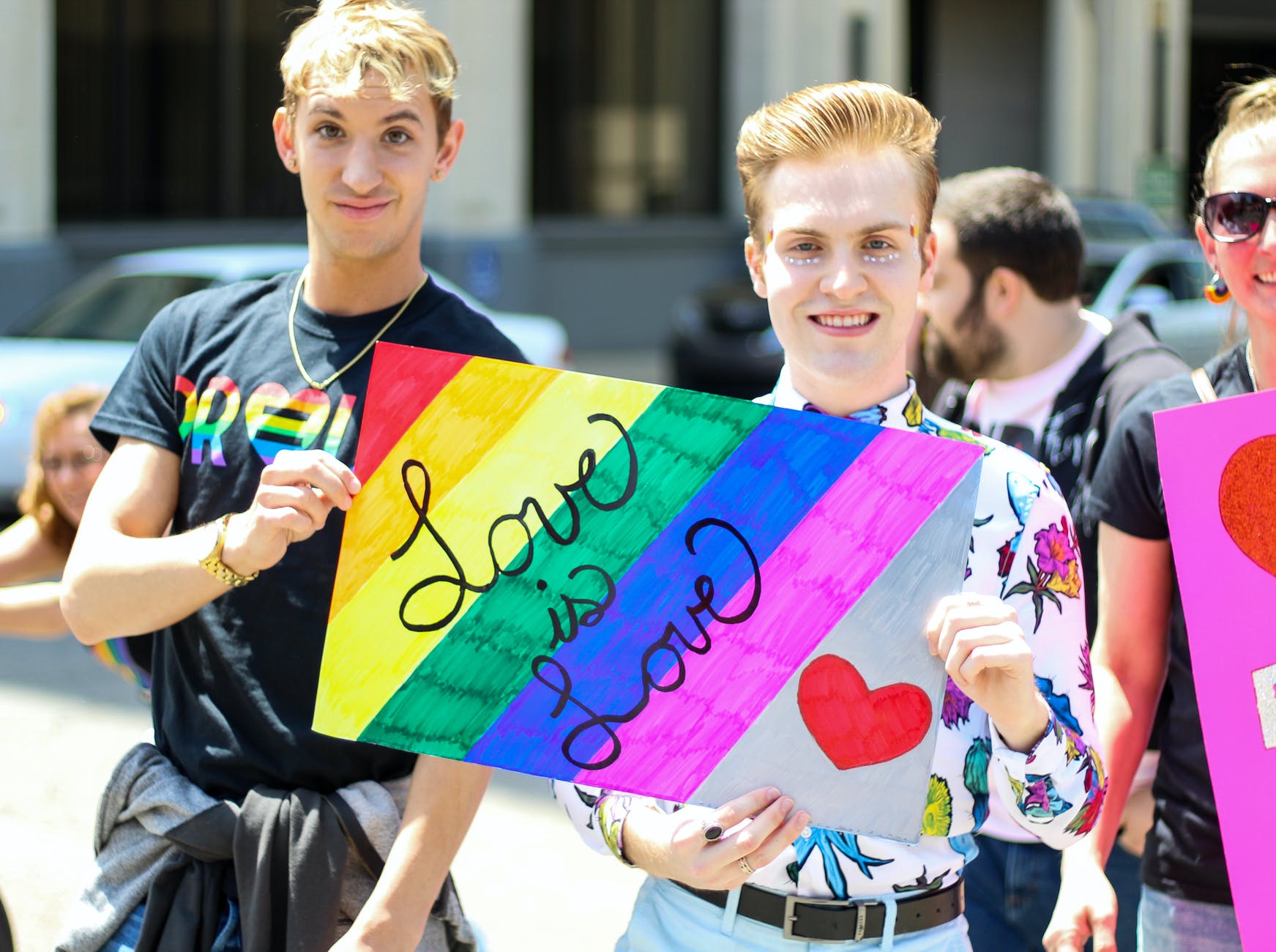 Two white, male-presenting people hold a "love is love" poster at a parade.