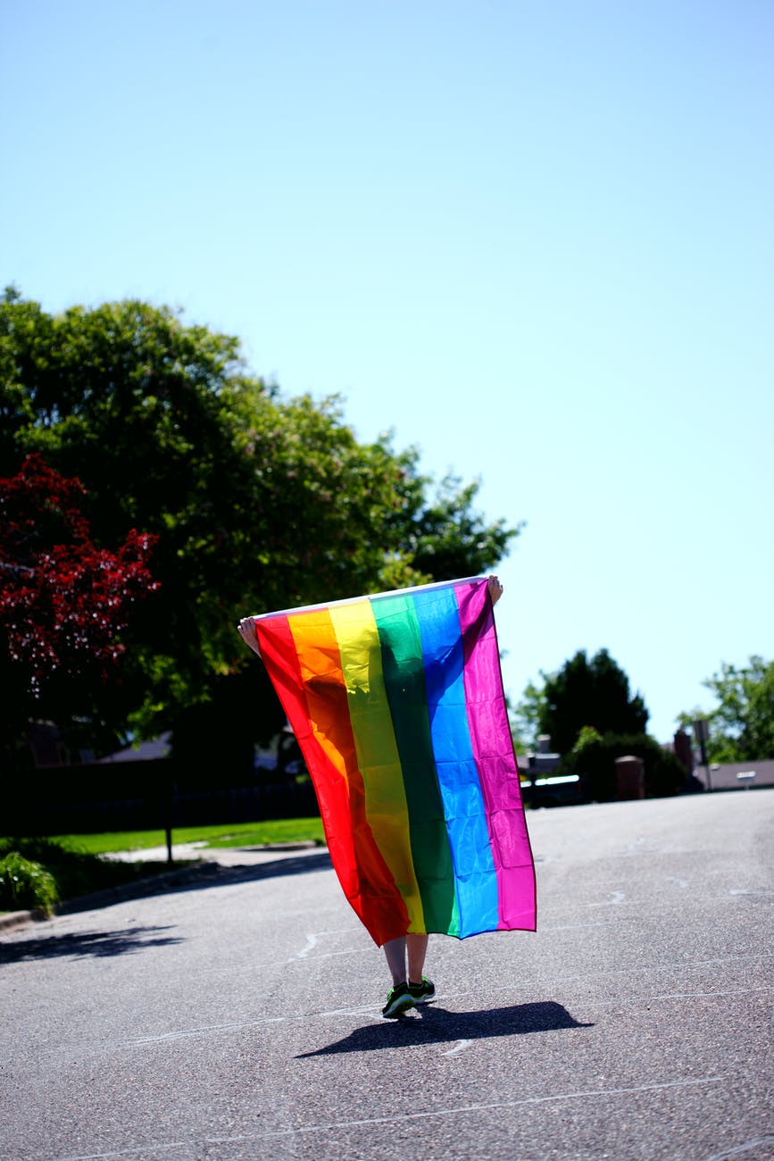 A person walks across a parking lot on a sunny day, a silhouette before the rainbow pride flag they carry behind them.